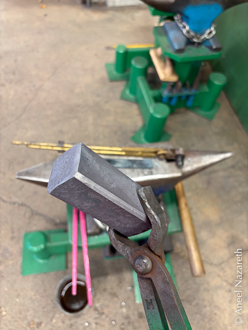 A hammer blank held in a pair of tongs. It's a rectangular prism with two square faces. The long edges are chamfered
